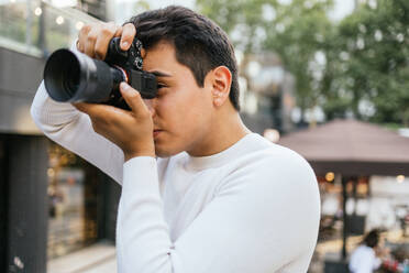 Caucasian young man wearing white t-shirt photographing with camera while standing against blurred background in city - ADSF47143