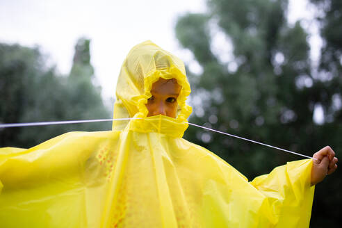 Unrecognizable child wearing yellow raincoat standing against blurred green trees in forest on rainy weather during daytime - ADSF47147