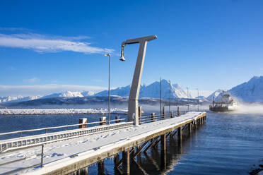 Snow covered long pier built on rippling sea with boat travelling against blue sky and mountains on sunny day in coastal area of Lofoten Islands Norway - ADSF47172