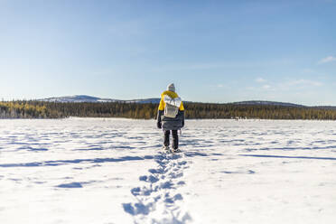 Back view of unrecognizable tourist in warm clothes with backpack walking on snowy field during hiking under blue sky Lapland Norway - ADSF47180