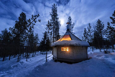 Exterior of small illuminated wooden cabin located on snowy forest in winter at night in Lapland Norway - ADSF47182