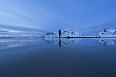 Distant view of unrecognizable tourist standing on shore near lake and snowy mountains under sky in winter Lofoten Islands - ADSF47185