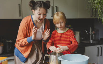 Blond girl breaking egg and sister cheering her in kitchen - IHF01692