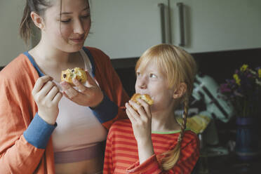 Blond girl eating muffins with elder sister in kitchen at home - IHF01703