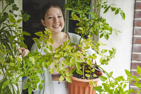 Lächelndes Teenager-Mädchen mit Topf-Tomatenpflanze auf dem Balkon - IHF01724