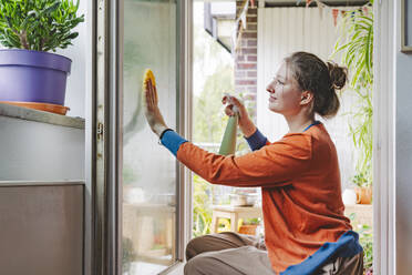 Teenage girl spraying water and cleaning door of balcony at home - IHF01734