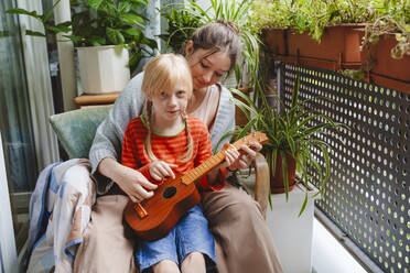 Teenage girl teaching sister how to play ukulele on balcony - IHF01743