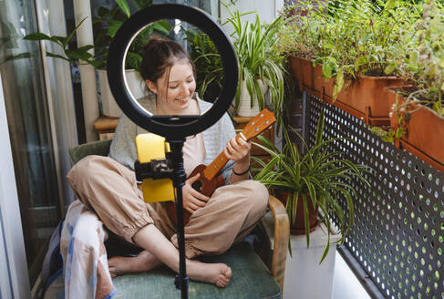 Influencerin spielt Ukulele und führt einen Vlog über ihr Smartphone auf dem Balkon - IHF01746