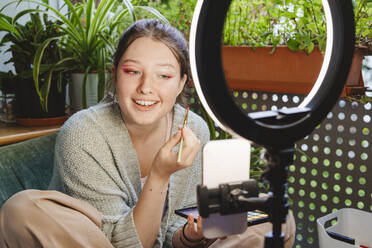 Smiling vlogger recording make-up video on smart phone using ring light - IHF01749