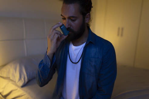 Man using inhaler sitting on bed at home - JPTF01321