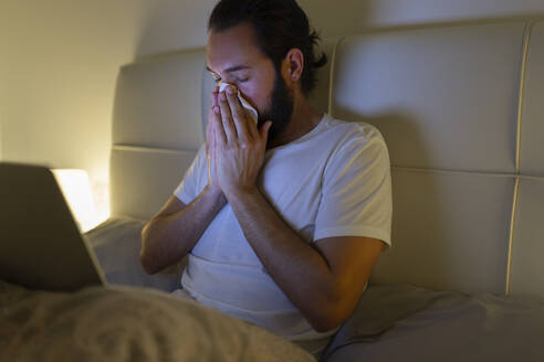 Man blowing nose and sitting with laptop on bed - JPTF01322