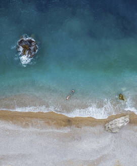 Luftaufnahme von zwei Männern, die in kristallklarem, von der Sonne beleuchtetem Wasser an einem leeren Strand schwimmen, Cala Moraig, Zona Encinas, Cumbre del Sol, Alicante, Spanien. - AAEF22089