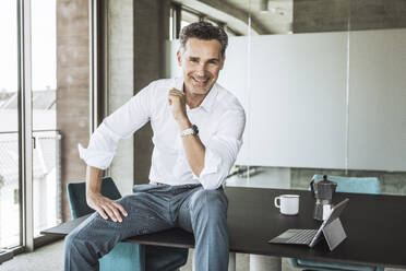 Happy businessman sitting on desk in office - UUF30354