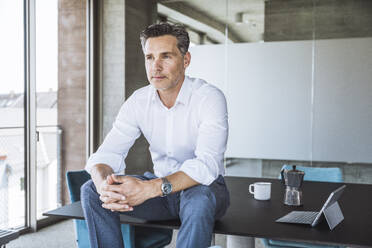 Thoughtful mature businessman with hands clasped sitting on desk - UUF30355