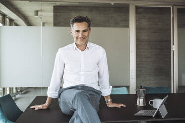 Smiling mature businessman sitting near tablet PC on desk - UUF30360