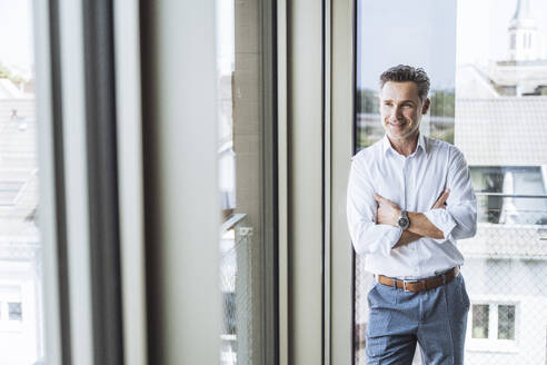 Lächelnder Geschäftsmann steht am Fenster im Büro - UUF30368