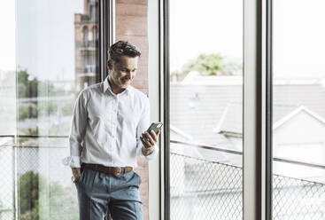 Lächelnder Geschäftsmann, der ein Smartphone in der Nähe eines Fensters im Büro benutzt - UUF30370