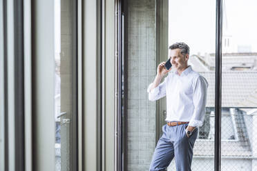 Smiling businessman talking on smart phone near window - UUF30371