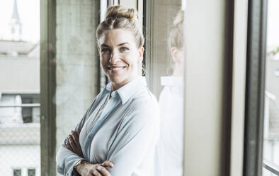 Happy businesswoman standing with arms crossed near window in office - UUF30394