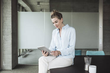 Lächelnde Geschäftsfrau sitzt auf dem Schreibtisch und benutzt einen Tablet-PC im Büro - UUF30397