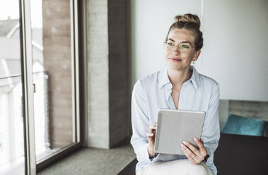 Thoughtful businesswoman wearing eyeglasses and sitting with tablet PC on desk - UUF30405