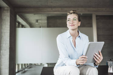 Smiling businesswoman wearing eyeglasses and sitting with tablet PC in office - UUF30408