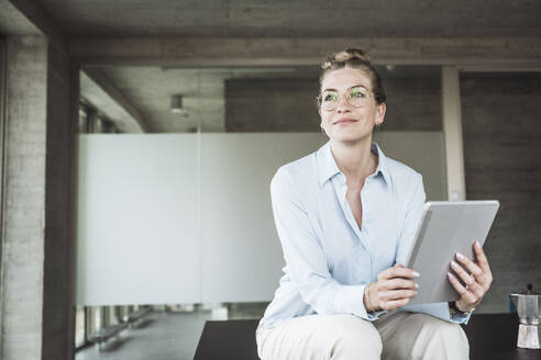 Lächelnde Geschäftsfrau, die eine Brille trägt und mit einem Tablet-PC im Büro sitzt - UUF30408