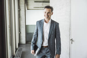 Happy mature businessman standing with tablet PC in office corridor - UUF30455