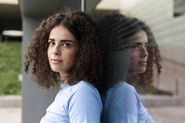 Junge Frau mit lockigem Haar lehnt an einem Glas - LMCF00560