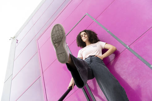Junge Frau mit hochgelegtem Bein vor einer rosa Tür stehend - LMCF00575