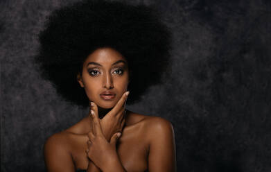 Seductive African American female model wearing black top with bare  shoulders leaning against the wall and closed eyes during photo session in  dark studio stock photo