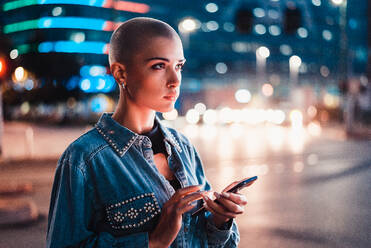 Pretty girl with stylish clothes holding smartphone outdoors in the evening, illuminated city on background - Concepts about technology, communication and lifestyle - DMDF05113