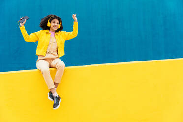 Junge glückliche Frau, die im Freien tanzt und Spaß hat. Teenager, der mit Smartphone und Kopfhörern in einem gelben und blauen modernen Stadtgebiet Musik hört - DMDF05146