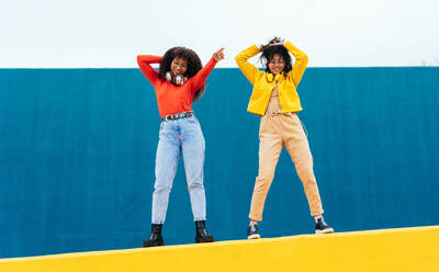 Young happy women dancing and having fun outdoor. Teenagers listening to music with smartphone and headphones in a yellow and blue modern urban area - DMDF05147