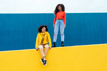 Young happy women posing on colored blue and yellow colored walls. Teenagers girl spending time together after school - DMDF05160