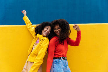 Young happy women dancing and having fun outdoor. Teenagers listening to music with smartphone and headphones in a yellow and blue modern urban area - DMDF05174