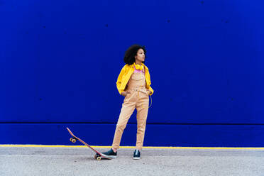 Young happy woman having fun outdoor with her skateboard. Teenager listening to music with smartphone and headphones in a yellow and blue modern urban area - DMDF05187