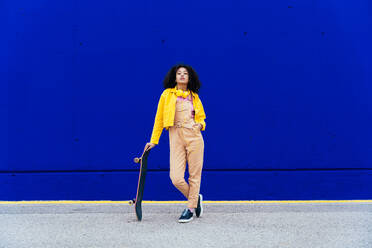 Young happy woman having fun outdoor with her skateboard. Teenager listening to music with smartphone and headphones in a yellow and blue modern urban area - DMDF05188