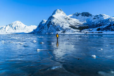 Distant back view of unrecognizable tourist standing on shore of frozen lake near snowy mountains against blue sky in winter Lofoten Islands - ADSF47187