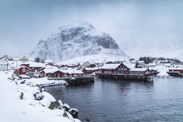 Amazing view of small settlement of houses located on shore of lake in snowy mountains against foggy sky in winter on Lofoten Islands in Norway - ADSF47190