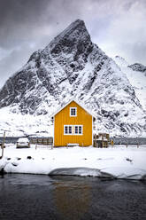 Spectacular landscape of small wooden yellow house located on frozen lake in snowy mountains against gray cloudy sky on Lofoten Islands in Norway - ADSF47192