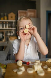 Cute playful Caucasian boy holding banana pieces in front of face while cutting fruits into slices on chopping board and cooking food in kitchen - ADSF47205