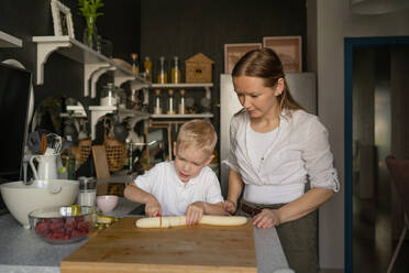 Mid adult Caucasian mother dressed in casuals looking at cute little son cutting banana into slices with knife on chopping board in kitchen - ADSF47206