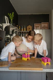 Caucasian twin sons with blonde hair looking curiously at mother pouring liquid in popsicle molds while making ice cream in kitchen during free time - ADSF47209