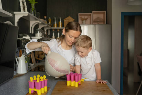 Caucasian son with blonde hair looking curiously at mother pouring liquid in popsicle molds while making ice cream in kitchen during free time - ADSF47210