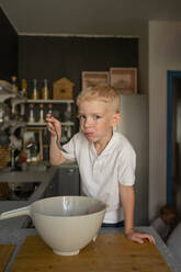 Caucasian cute boy with blonde hair standing trialling the bowl preparation of strawberries while cooking food in kitchen at home - ADSF47212