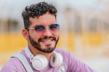 Smiling bearded male in casual clothes and eyeglasses with headphone sitting on stone bench while looking at camera and listening to music - ADSF47225