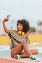 Positive young curly haired female in trendy clothes and earphones sitting on stone bench with mobile phone while taking selfie against blurred background - ADSF47226