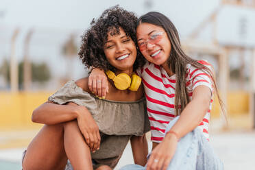 Laughing multiracial young girlfriends in trendy clothes embracing and looking at camera while sitting on pavement against blurred urban background - ADSF47227