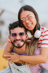 Cheerful young bearded man in sunglasses smiling while hugging girlfriend and looking at camera on blurred background of street with headset - ADSF47229